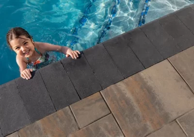 Smiling child holding onto pool edge, wearing a swimsuit in bright blue water, enjoying the beautifully designed hardscaping by Solid Brick Contracting.