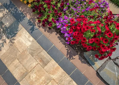 Red and purple flowers border a beautiful hardscaping stone pathway with tan, brown, and dark gray tiles in sunlight—an elegant landscape crafted by Solid Brick Contracting, MD.