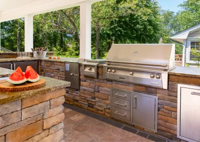 Outdoor kitchen with stainless steel grill, stone counters, and sliced watermelon on a cutting board—perfect for enjoying outdoor hardscaping and landscaping in MD.