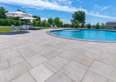 A sunny poolside patio in Aberdeen, MD, with expert hardscaping features, lounge chairs, umbrellas, and a clear blue swimming pool surrounded by lush greenery.