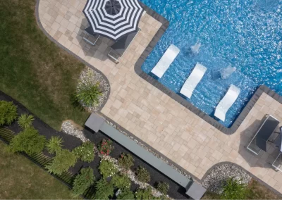 Aerial view of a pool with three lounge chairs, umbrella, patio, garden, and hardscaping features enhancing the grass area.