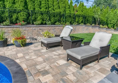 Two cushioned lounge chairs by a pool on a stone patio, with lush plants and a green hedge in the background—an inviting hardscaping feature designed for relaxing outdoor moments.