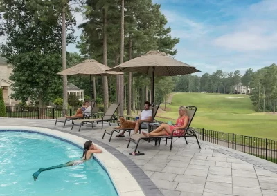 People relaxing by a pool with lounge chairs and umbrellas, enjoying expertly designed hardscaping and scenic views of the green golf course and trees in Aberdeen, MD.