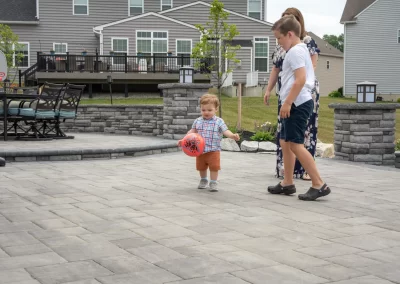 A toddler with a red ball walks on a hardscaping patio, followed by a woman and an older child in a suburban Aberdeen backyard.