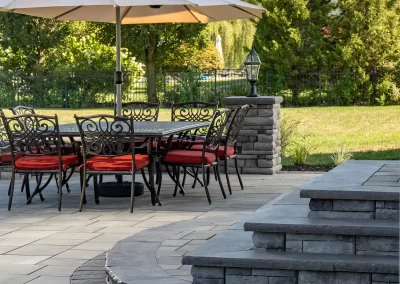 Outdoor patio with a dining table, chairs, red cushions, large umbrella, stone steps, and green trees in the background—expertly crafted by Solid Brick Contracting for exceptional hardscaping and beautiful landscaping.