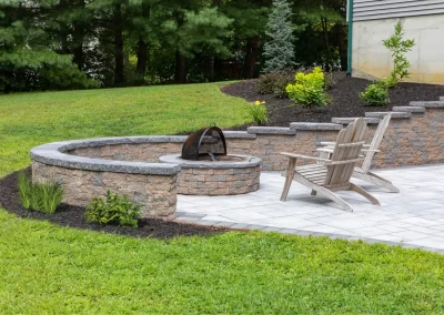 Two wooden chairs by a stone fire pit on a patio, crafted by Solid Brick Contracting, with beautiful landscaping and a lush MD garden lawn in the background.