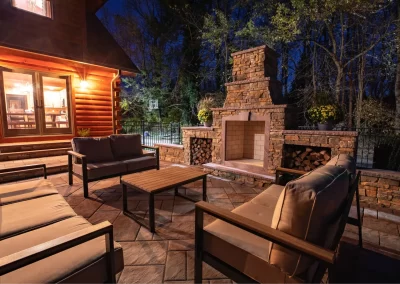 Cozy patio at night with cushioned seating, wooden table, and a stone outdoor fireplace beside a log cabin, showcasing expert landscaping by Solid Brick Contracting, MD.
