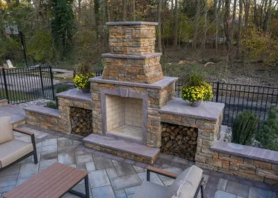 Stone outdoor fireplace with yellow flowers, wood storage, and patio seating highlights the hardscaping expertise of Solid Brick Contracting in a beautifully landscaped, fenced backyard.