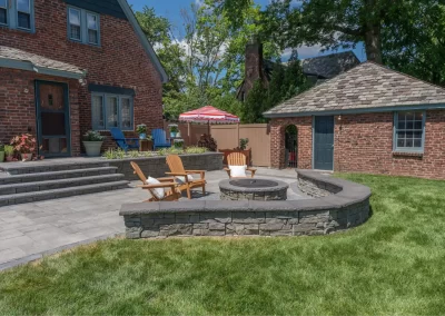 Stone patio with fire pit, wooden chairs, and outdoor seating area in a grassy backyard next to a brick house—expertly designed by Solid Brick Contracting for standout landscaping in Aberdeen.