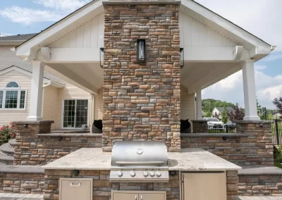 Experience exceptional hardscaping with this outdoor stone kitchen by Solid Brick Contracting, MD—featuring a built-in grill and chimney beneath a white covered patio on a sunny day.
