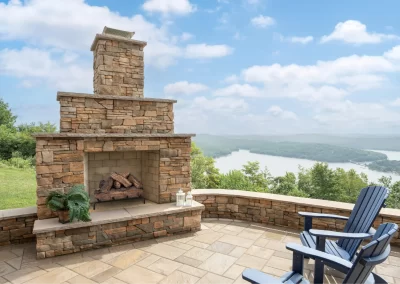 Stone outdoor fireplace on a patio with blue chairs, overlooking a scenic lake and green hills—perfect for relaxing in Aberdeen, MD and enhancing your landscaping.