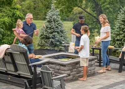 A family in MD gathers around a backyard fire pit, enjoying roasted marshmallows and quality time together, surrounded by beautiful hardscaping and landscaping features.