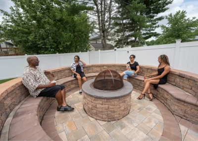 Four people sit and talk around a circular backyard fire pit on a stone patio, showcasing expert hardscaping by Solid Brick Contracting, surrounded by trees and a fence in Aberdeen.