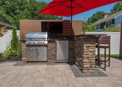 Outdoor patio kitchen with a grill, mini fridge, TV, bar seating, and a large red umbrella overhead—expertly designed by Solid Brick Contracting to blend stylish hardscaping with modern outdoor living.