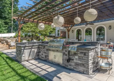 Outdoor stone kitchen with grill under a wooden pergola and hanging lanterns, surrounded by green grass—an inviting Aberdeen hardscaping project by Solid Brick Contracting.