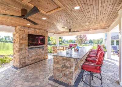 Covered outdoor kitchen with stone bar, red barstools, TV, ceiling fans, and a view of expertly designed landscaping featuring a lush lawn and trees.