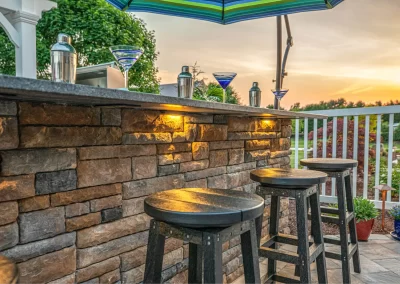 Outdoor stone bar with stools, martini glasses, and shakers at sunset under a striped umbrella—perfect for entertaining in your Aberdeen backyard. Elevate your landscaping with expert touches from Solid Brick Contracting.