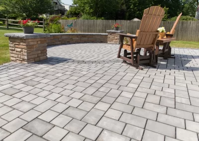 Two wooden chairs and small tables on a paved patio showcase beautiful hardscaping in a sunny MD backyard, complete with a stone wall and lush landscaping.