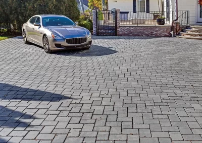 A silver car is parked on a spacious driveway made of gray interlocking pavers by Solid Brick Contracting, showcasing expert hardscaping in front of a house.