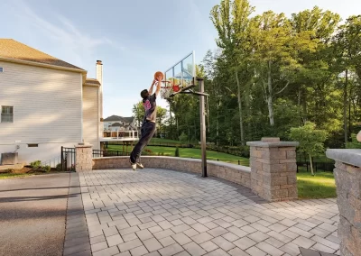 Person jumping to dunk a basketball on an outdoor hoop in a sunny backyard, surrounded by beautiful landscaping and hardscaping features designed by Solid Brick Contracting, with trees and houses nearby.