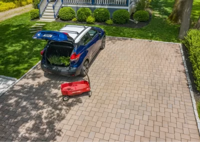 A blue car with an open trunk and a red wagon sits on a brick driveway near a house in MD, surrounded by green shrubs and well-done landscaping.