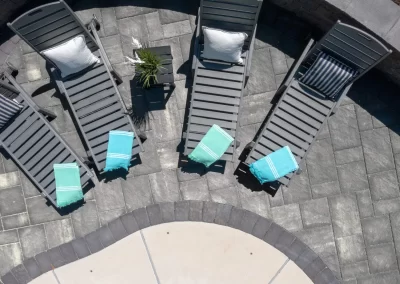 Four gray lounge chairs with pillows and towels sit on a stone patio beside a pool, viewed from above at a beautiful Aberdeen, MD residence crafted by Solid Brick Contracting.