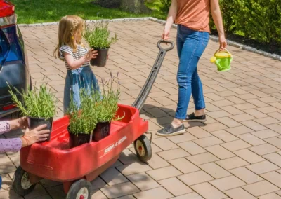 A woman pulls a red wagon with plants while a young girl carries a potted plant on a sunny driveway in Aberdeen, MD.