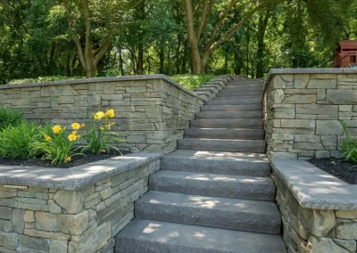 Stone steps with flower beds and yellow flowers create inviting hardscaping, leading up to a wooded area with lush green trees—perfect for beautiful MD landscaping.