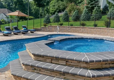 Tiered stone hot tub attached to a swimming pool, with lounge chairs and umbrella set in expertly designed Aberdeen backyard landscaping.