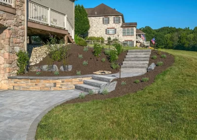 Stone steps and a retaining wall by Solid Brick Contracting enhance this landscaped Aberdeen, MD backyard, with mulch, lush plants, and a large house in the background.