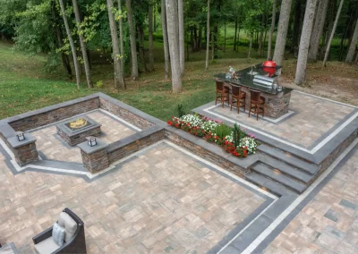 Outdoor patio in Aberdeen, MD with tiered stone seating, fire pit, bar, grill, barstools, and surrounded by trees—expertly crafted by Solid Brick Contracting.