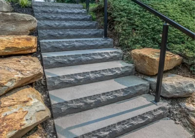 Stone outdoor steps with black metal railings, bordered by rocks and greenery, lead uphill—showcasing expert hardscaping by Solid Brick Contracting, MD.