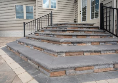 Stone patio steps with black railings enhance the hardscaping design, leading to a house with beige siding and glass door in MD.