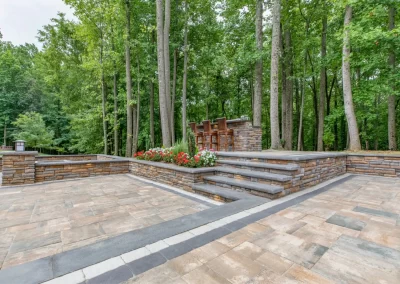 Paved patio with stone walls, stairs, flowers, and a bar area, expertly crafted by Solid Brick Contracting in Aberdeen, MD—surrounded by tall trees in a peaceful forest setting.