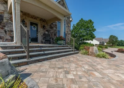 Stone porch steps with railings lead to a house with stone walls in Aberdeen, surrounded by expert hardscaping, lush landscaping, and a paved patio.