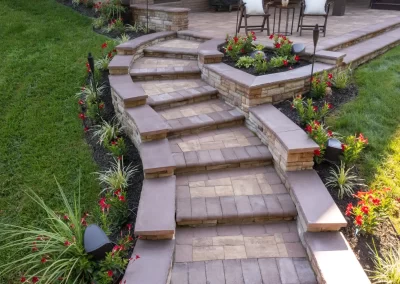 Stone steps with flower beds and decorative lighting, expertly crafted by Solid Brick Contracting, lead up to a patio with chairs on a grassy lawn—an inviting hardscaping feature perfect for any MD home.