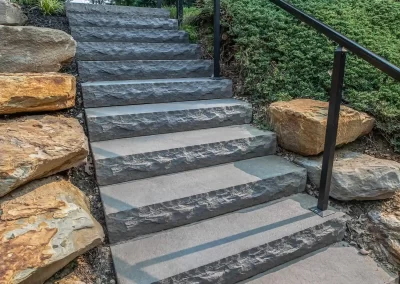 Stone stairs with black railing, bordered by large rocks and green shrubs, lead up a grassy hill—an elegant hardscaping project crafted by Solid Brick Contracting in Aberdeen.