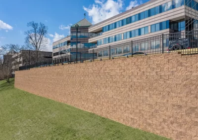 A tall retaining wall, expertly built by Solid Brick Contracting, with a black fence in front of a modern office building on a grassy Aberdeen slope, showcases quality hardscaping.