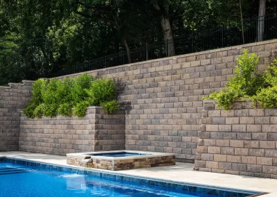 A pool with a small waterfall, set against a tall stone retaining wall topped with green shrubs—showcasing expert landscaping and hardscaping by Solid Brick Contracting.