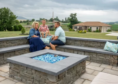 Three people sit and talk around a stone firepit on a beautifully landscaped patio by Solid Brick Contracting, with a scenic rural Aberdeen background.