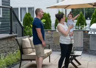 A man and woman stand outside on a patio, possibly designed by Solid Brick Contracting; the woman holds a smiling toddler who is pointing upwards, enjoying the professionally crafted hardscaping in MD.