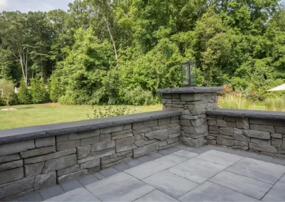 Stone patio with a short stone wall, overlooking a green lawn and dense trees in the background—perfect for enjoying expertly crafted landscaping in Aberdeen, MD.