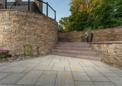 Stone patio with curved stone wall, stairs, potted plants, and trees in the background at sunset—a stunning hardscaping project by Solid Brick Contracting in Aberdeen.
