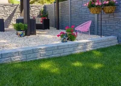 A modern backyard in MD with lush green grass, stylish hardscaping, a stone patio, pink chair, and potted flowers set against a sleek gray stone wall.