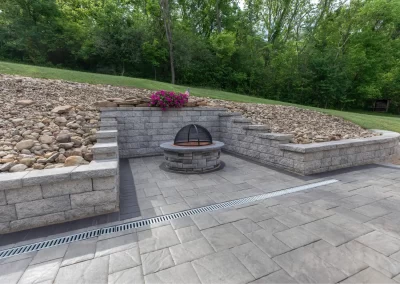 Stone patio with a round fire pit, retaining walls, and a flower planter; trees in the background. Expert hardscaping by Solid Brick Contracting brings your MD outdoor space to life.
