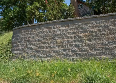 A stone retaining wall by Solid Brick Contracting with grass in front, trees, and part of a house visible above the wall—a fine example of hardscaping craftsmanship in Aberdeen.