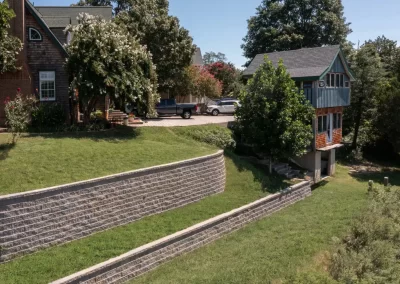 Two houses in Aberdeen, MD feature a sloped yard with stone retaining walls, green grass, and trees on a sunny day—showcasing the quality work of Solid Brick Contracting.