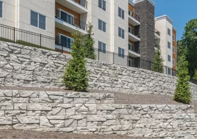 Stone retaining walls and young trees enhance the landscaping in front of a modern multi-story apartment building on a sunny day, expertly crafted by Solid Brick Contracting, MD.