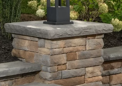 A black outdoor lantern sits on a stone pillar next to stone steps and green bushes, creating a welcoming entrance. Expertly crafted by Solid Brick Contracting in Aberdeen, MD.