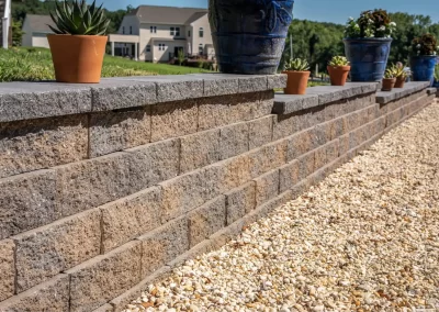 Stone retaining wall with potted plants on top, alongside a gravel path and houses in the background—expert landscaping by Solid Brick Contracting, MD.
